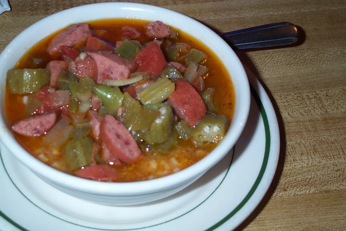A white bowl of gumbo with sausage, okra and rice on a wooden table