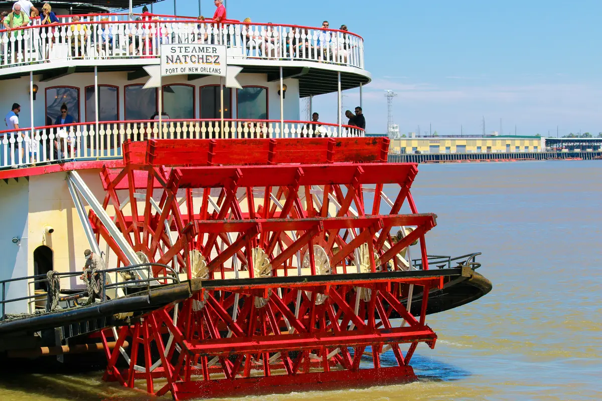 The red sternwheel of the Steamboat Natchez with the name board and deck rails above it and the brown river beside it