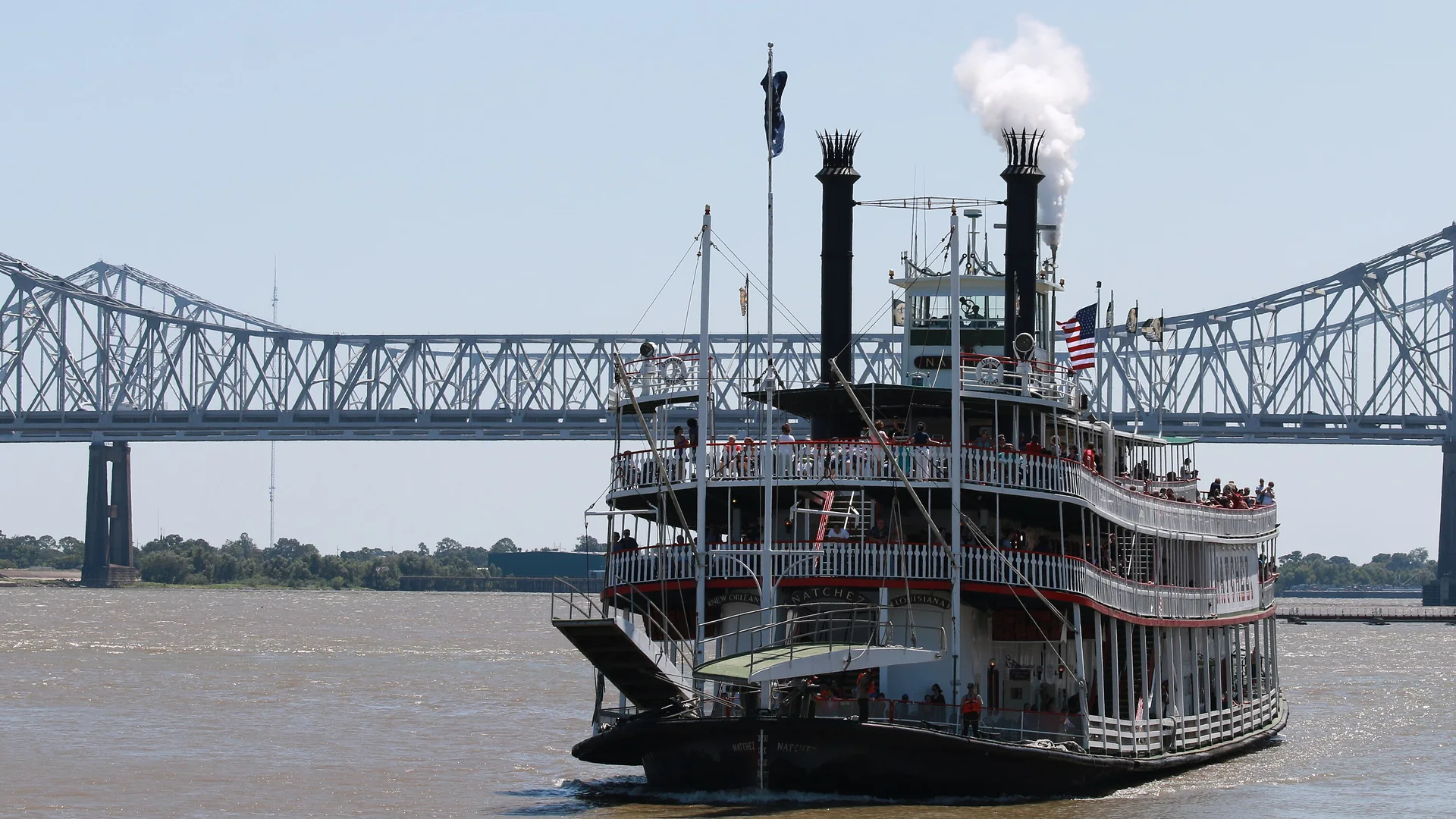 The Steamboat Natchez seen from the front on the Mississippi, with two black chimneys, a plume of steam and a steel bridge behind her.